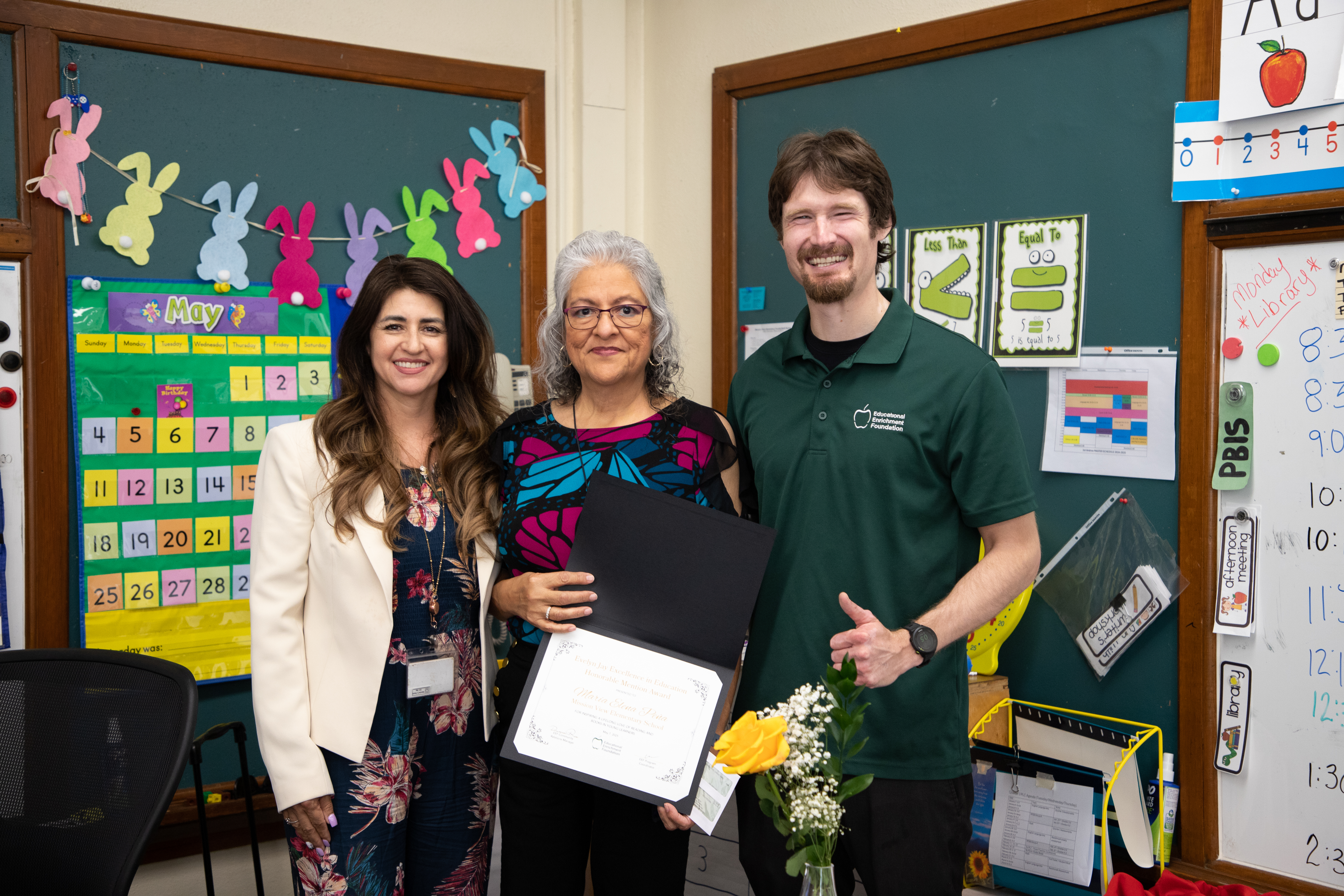 A woman with grey hair shows off her award certificate, next to a woman and man