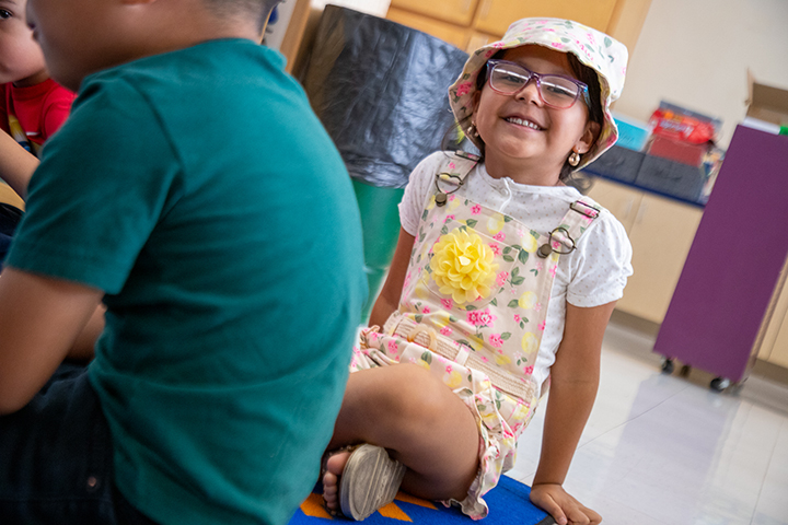 A little girl in glasses and a floral hat and shortalls smiles while sitting on the classroom floor