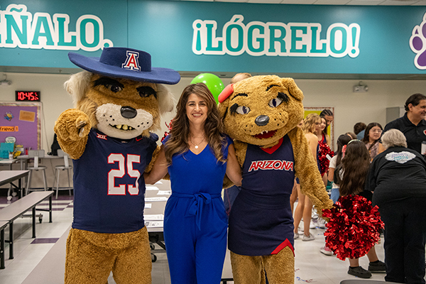 A wildcat mascot in a jersey and hat and a wildcat mascot in a cheerleader dress pose with a woman in a blue jumpsuit