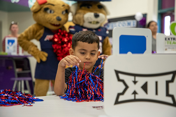 Two Wildcat mascots stand behind a boy as he plays with a blue and red pom pom