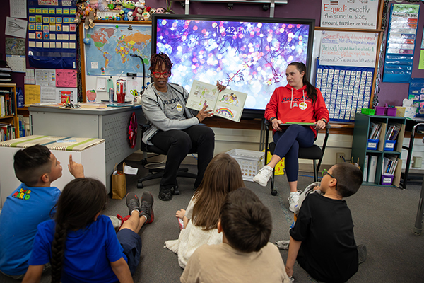 A woman in a gray hoodie reads a picture book to kids sitting on the floor, as a woman in a red hoodie looks on