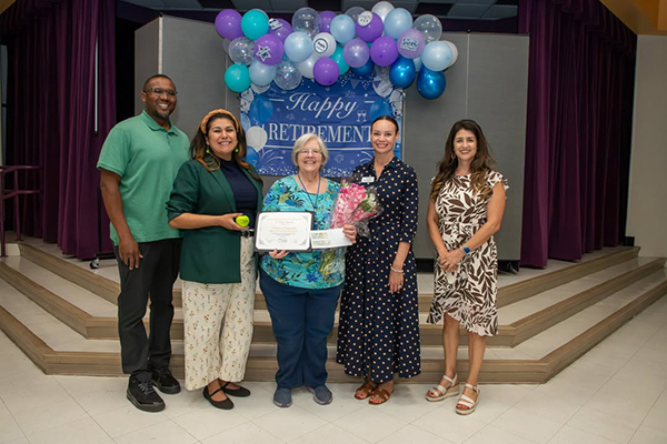 A woman, center, proudly holds up an award and flowers, surrounded by her colleagues and principal