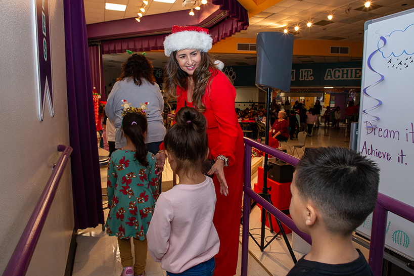 A woman in a red dress and Santa hat guides students up onto the stage