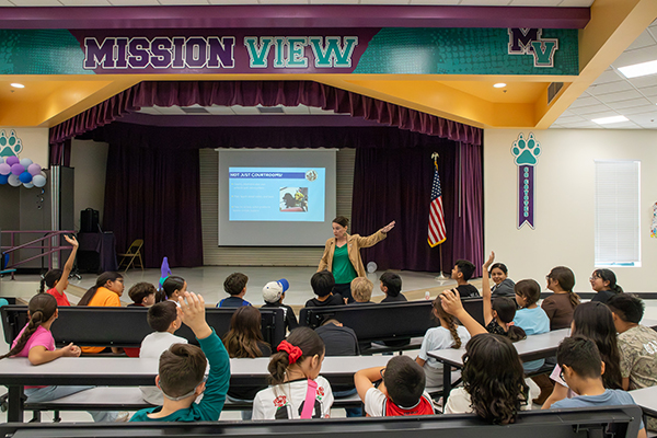 Students raise their hands in the audience as a woman talks to them