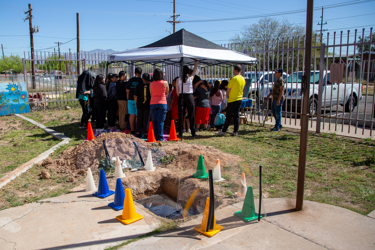 Students and staff stand under a tent with the hole where the time capsule will be buried in the foreground
